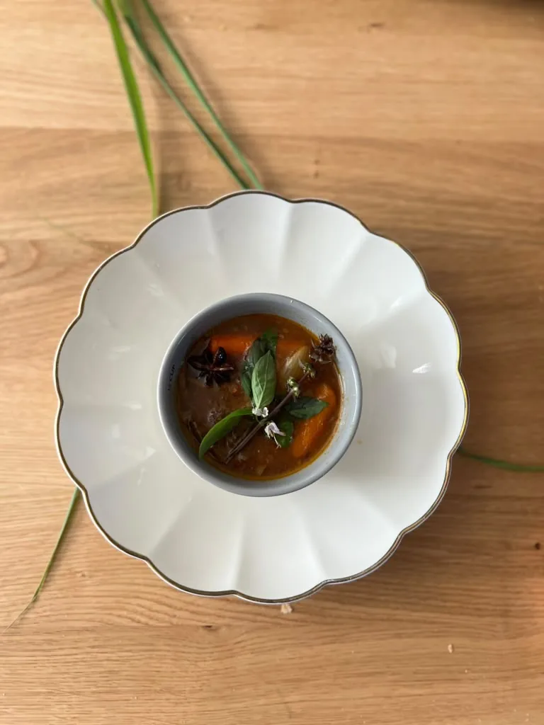 A small bowl of dark broth finished with fresh herbs, shot from above on a scalloped plate