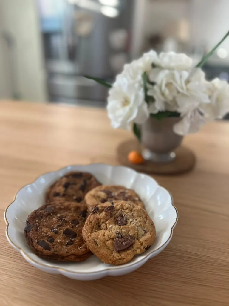 Three chocolate chip cookies on a scalloped plate beside a vase of white carnations